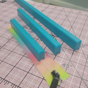 May include: Three turquoise zipper guides and a rainbow gradient zipper on a pink cutting mat. The zipper is open, revealing the teeth and the zipper pull. The guides are rectangular and appear to be made of plastic.
