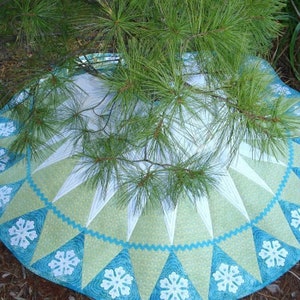 May include: A circular, quilted table runner with a snowflake design in shades of blue, green, and white. The runner is decorated with a scalloped edge and a repeating pattern of snowflakes.