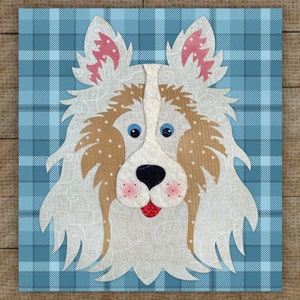 May include: A fabric art piece featuring a dog's face with a white and tan coat, blue eyes, and a black nose. The dog has pink accents on its ears and cheeks. The background is a blue plaid pattern.