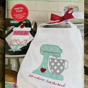 Puede incluir: Un delantal blanco con un diseño de batidora turquesa y la frase "life is short, lick the bowl". Un lazo rojo adorna la parte superior. La portada de un patrón de bordado Kimberbell Classic Vol. 6 es visible.