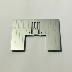 Puede incluir: Una placa plateada de máquina de coser con marcas grabadas para mediciones. La placa tiene líneas verticales y números, incluyendo 10, 20, 30 y 40, con medidas fraccionarias en pulgadas correspondientes. Cuenta con un recorte rectangular y dos pequeños agujeros.