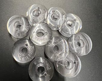 May include: A collection of clear plastic sewing machine bobbins. Each bobbin has a circular design with a central hole and the word "SINGER" imprinted on the surface. The bobbins are arranged on a dark surface.