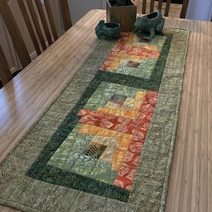 Pode incluir: Um corredor de mesa de patchwork verde, laranja e amarelo com um design geométrico. O corredor tem uma borda verde e está sobre uma mesa de madeira.