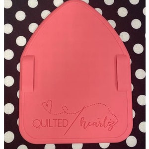 Könnte beinhalten: Eine rosa Silikon-Bügelmatte mit dem Text "QUILTED heartz" und einem stilisierten Nadel- und Faden-Design.