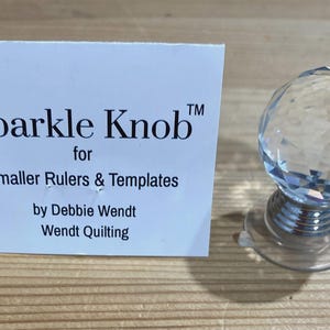 Könnte beinhalten: Ein klarer, facettierter Kristallknopf mit silberfarbenem Sockel und Saugnapf, neben einer weißen Karte mit dem Text "Sparkle Knob" für kleinere Lineale und Schablonen von Debbie Wendt, Wendt Quilting.