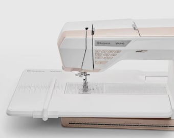 May include: A white and gold Husqvarna Viking sewing machine. The machine has a white body with gold accents and a control panel with multiple buttons. A white extension table with measurement markings is attached to the machine.