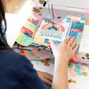 Puede incluir: Primer plano de una persona usando una máquina de coser para bordar una tela colorida con el texto "Choose Kindness". La tela está rodeada de confeti.