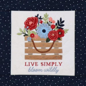 Puede incluir: Diseño floral bordado con una cesta de madera llena de flores rojas, azules y rosas. El texto "LIVE SIMPLY bloom wildly" está bordado debajo de la cesta. El diseño está sobre un fondo blanco con un borde azul marino de lunares.