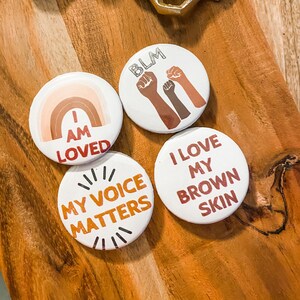 May include: Four white round buttons with text and graphics. One button says "I AM LOVED" with a rainbow design. Another says "BLM" with raised fists. The third says "MY VOICE MATTERS". The last says "I LOVE MY BROWN SKIN".