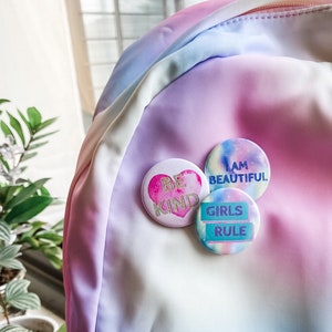 May include: Three round pinback buttons with positive messages are pinned to a pastel rainbow backpack. The buttons read "BE KIND", "I AM BEAUTIFUL", and "GIRLS RULE". The backpack has a zipper and is made of a soft, silky material.