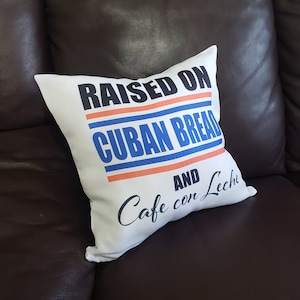 Puede incluir: Almohada blanca con el texto "RAISED ON CUBAN BREAD AND Cafe con Leche" en letras negras y azules. La almohada está en un sofá de cuero marrón.