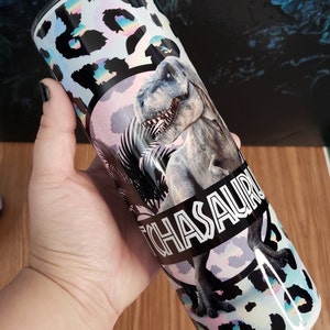 May include: A stainless steel tumbler featuring a T-Rex dinosaur design with the word "T-CHASAURUS" in white. The tumbler has a leopard print pattern in shades of blue, pink, and black.