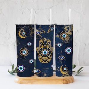 May include: Three stainless steel tumblers with lids and straws. Each tumbler features a different design with a blue background and gold accents. The designs include a crescent moon, a hamsa hand, and an evil eye.