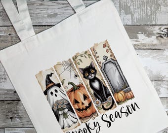 Op de afbeelding: Witte canvas draagtas met een Halloween-thema ontwerp. Het ontwerp bevat een spook, pompoen, zwarte kat en grafsteen. De woorden "Spooky Season" staan eronder in een scriptlettertype.