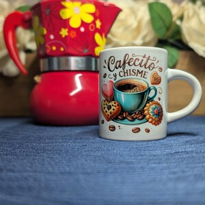 May include: A white coffee mug with the words "Cafecito y Chisme" and a coffee cup design, accompanied by heart and cookie illustrations. A red coffee maker with yellow floral designs is in the background.