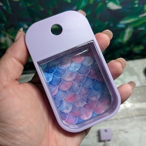 May include: A rectangular, light purple plastic hand sanitizer holder featuring a clear front window. The insert inside displays a repeating pattern of overlapping scales in shades of blue, purple, and pink with a subtle, speckled galaxy-like effect.