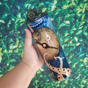 Puede incluir: Un vaso de acero inoxidable azul y dorado con un diseño de esfera de reloj y tentáculos de pulpo. El vaso tiene una tapa de metal dorado con un engranaje y otros adornos de estilo steampunk.