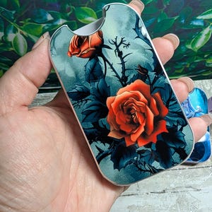 May include: A decorative case featuring a vibrant orange rose design against a teal background. The case has a silver trim and a curved shape. The floral artwork includes dark blue leaves and thorny stems.