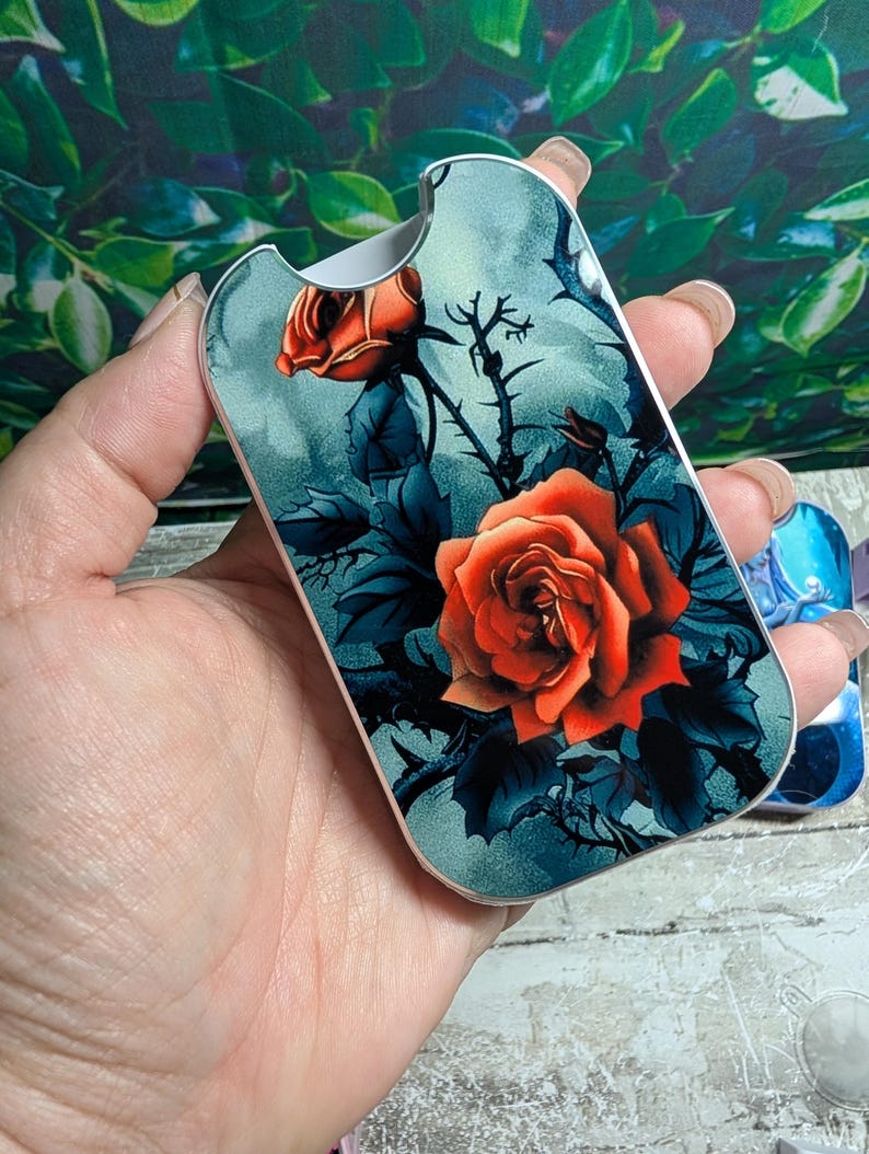 May include: A decorative case featuring a vibrant orange rose design against a teal background. The case has a silver trim and a curved shape. The floral artwork includes dark blue leaves and thorny stems.