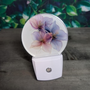 May include: A white night light with a round, translucent front panel featuring a watercolor-style floral design of pink and purple flowers. The light is on and illuminating the flowers.