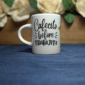 May include: White ceramic mug with a curved handle. The mug features the text "Cafecito before TRABAJITO" in black script. The mug is set against a blue background with a blurred floral arrangement.