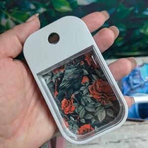 May include: A white, rectangular hand sanitizer bottle with a clear window displaying a floral design. The design features a black raven amidst red roses and green leaves on a black background. The bottle has a hole at the top for attachment.