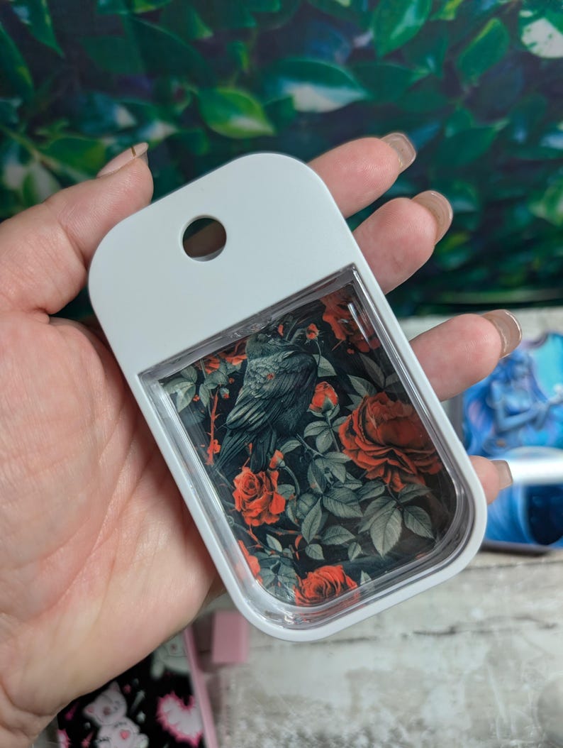 May include: A white, rectangular hand sanitizer bottle with a clear window displaying a floral design. The design features a black raven amidst red roses and green leaves on a black background. The bottle has a hole at the top for attachment.