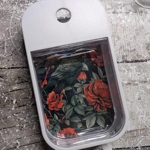 May include: A white rectangular tray with a rounded top and a hole. The tray's interior features a floral design with red roses, green leaves, and a dark bird against a black background. The tray is likely used for holding small items.