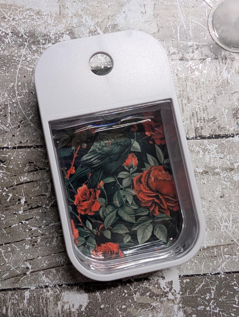 May include: A white rectangular tray with a rounded top and a hole. The tray's interior features a floral design with red roses, green leaves, and a dark bird against a black background. The tray is likely used for holding small items.