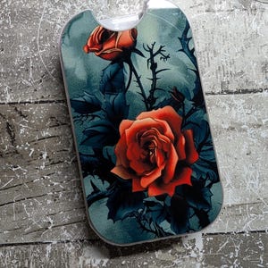 May include: A phone case featuring a vibrant red rose design against a teal background. The case has a curved top and a dark floral pattern with thorns and leaves. The rose is the focal point, with detailed petals.