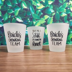 May include: Three frosted shot glasses with black text. Two glasses read "Bride's DRINKING TEAM", and the center glass says "BUY ME A Shot I'M TYING THE knot". The glasses are on a wooden surface with a green leafy background.