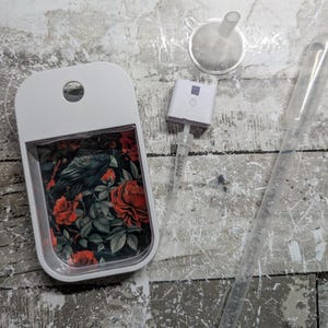 May include: A white, rectangular perfume bottle with a floral design featuring red roses and a raven. Includes a small funnel, a spray nozzle, and a clear pipette. The bottle has a hole at the top for a lanyard.