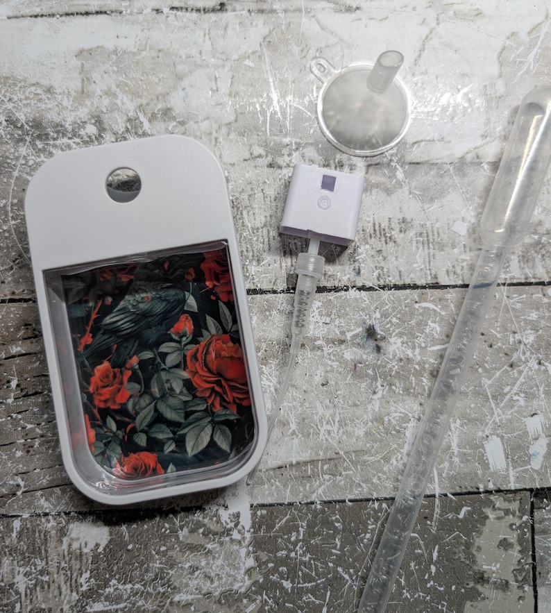 May include: A white, rectangular perfume bottle with a floral design featuring red roses and a raven. Includes a small funnel, a spray nozzle, and a clear pipette. The bottle has a hole at the top for a lanyard.
