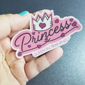 Könnte beinhalten: Rosafarbener Aufkleber mit dem Wort "Princess" in schwarzer Schrift mit einer Krone und Herzen. Der Text "Queen in Training" befindet sich unter dem Wort "Princess".