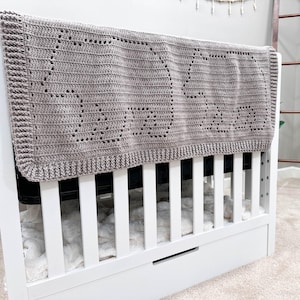 May include: A gray crocheted baby blanket with a decorative pattern hangs over a white crib with a white mattress and a white drawer.
