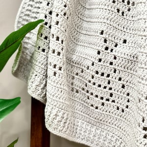 May include: A close-up of a crocheted throw blanket in a light gray color. The blanket has a delicate, openwork pattern.