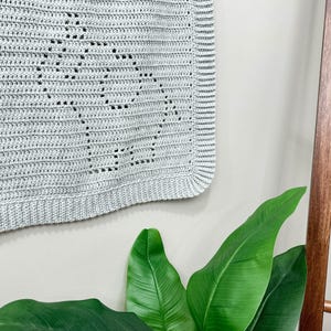 May include: A light gray crocheted baby blanket with a delicate pattern. The blanket is hanging on a wall next to a wooden ladder.