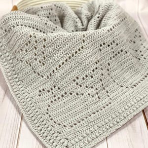 May include: A light gray crocheted blanket with a textured pattern and a decorative border. The blanket is folded and placed in a basket. The handmade blanket is a soft, cozy item.