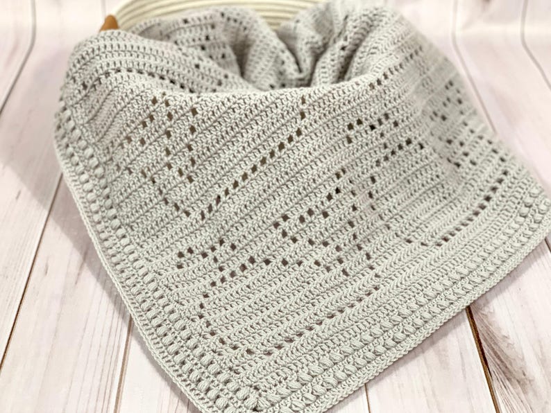 May include: A light gray crocheted blanket with a textured pattern and a decorative border. The blanket is folded and placed in a basket. The handmade blanket is a soft, cozy item.
