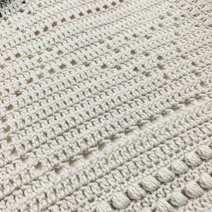 May include: Close-up of a handmade crochet blanket in shades of gray. The blanket features a textured pattern with various stitch designs, including openwork and bobble stitches. The soft, neutral colors and intricate details suggest a cozy, handcrafted item.