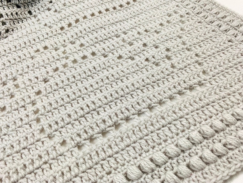 May include: Close-up of a handmade crochet blanket in shades of gray. The blanket features a textured pattern with various stitch designs, including openwork and bobble stitches. The soft, neutral colors and intricate details suggest a cozy, handcrafted item.