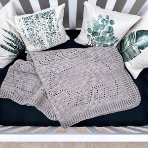 K&ouml;nnte beinhalten: Eine graue geh&auml;kelte Babydecke mit einem zarten Muster, ausgelegt auf einer schwarzen Babybettmatratze mit drei wei&szlig;en Kissen mit gr&uuml;nen botanischen Drucken.