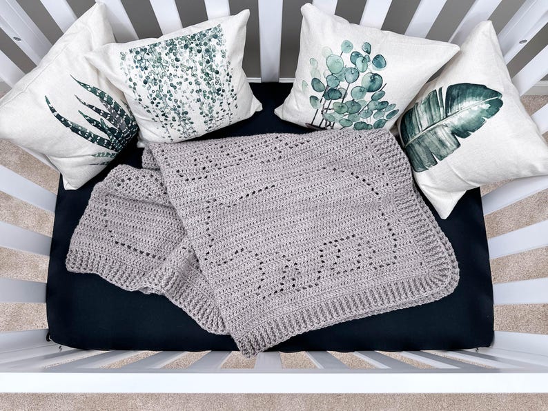 K&ouml;nnte beinhalten: Eine graue geh&auml;kelte Babydecke mit einem zarten Muster, ausgelegt auf einer schwarzen Babybettmatratze mit drei wei&szlig;en Kissen mit gr&uuml;nen botanischen Drucken.