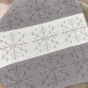 May include: A handmade crochet blanket in a square shape. The blanket features a central white stripe with a snowflake design, bordered by gray crochet. The blanket is made of soft yarn.