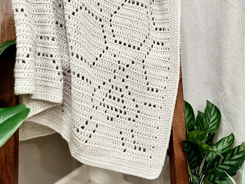 May include: A crocheted throw blanket in a light beige color with a geometric pattern. The blanket is draped over a wooden chair.