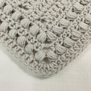 May include: Close-up of a crocheted item in a light gray color. The crochet pattern features a textured, raised design with a series of small, rounded bumps. The item is placed on a white, textured surface, with the corner of the crocheted piece visible.