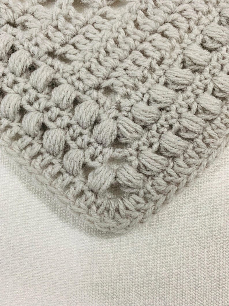 May include: Close-up of a crocheted item in a light gray color. The crochet pattern features a textured, raised design with a series of small, rounded bumps. The item is placed on a white, textured surface, with the corner of the crocheted piece visible.