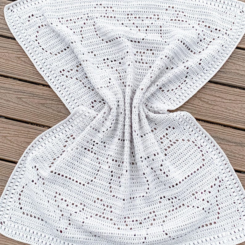May include: A white, crocheted blanket with a heart pattern. The blanket is folded and draped over a wooden surface. The crochet work is detailed, with open spaces creating the heart design.