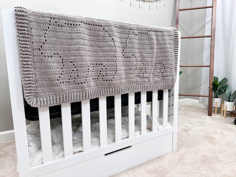 K&ouml;nnte beinhalten: Eine graue geh&auml;kelte Babydecke mit einem dekorativen Muster h&auml;ngt &uuml;ber einem wei&szlig;en Babybett mit einer wei&szlig;en Matratze und einer wei&szlig;en Schublade.
