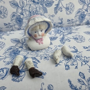 May include: A porcelain doll head and torso bust featuring a bonnet with pink and blue trim, accompanied by two separate porcelain legs with brown boots and two separate porcelain arms, all rendered in a white finish with painted facial details.